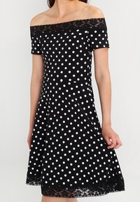 Svart off-shoulder-klänning med vita prickar, spetsdetaljer, figursydd överdel och utsvängd kjol. Korta ärmar och slät yta.