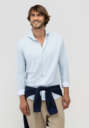 Hombre sonriente vistiendo una camisa de botones a rayas azul claro, pantalones beige y un suéter azul oscuro atado alrededor de su cintura sobre un fondo liso.