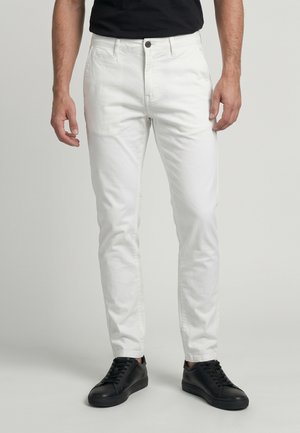 Homme portant un pantalon blanc ajusté et des baskets noires, debout devant un fond clair uni.