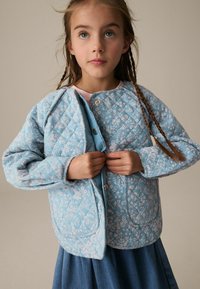 Giacca trapuntata blu chiaro con motivo floreale, chiusura frontale a pressione e due tasche, indossata sopra una maglietta rosa, abbinata a una gonna di jeans.