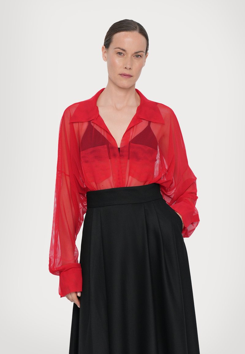 Blusa trasparente rossa a maniche lunghe con colletto, sovrapposta a una gonna nera a vita alta in tessuto morbido e vestibilità comoda.