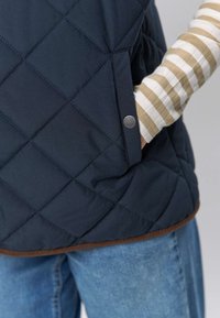 Marineblå quiltet vest med diamantmønster, med metallisk trykknapp og brun kant, båret over en stripete langermet skjorte.