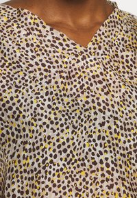 Lekki top z dekoltem w kształcie V, w beżowej bazie ozdobionej nieregularnymi łatakami w kolorze brązowym i żółtym, przypominającymi lamparcie cętki.