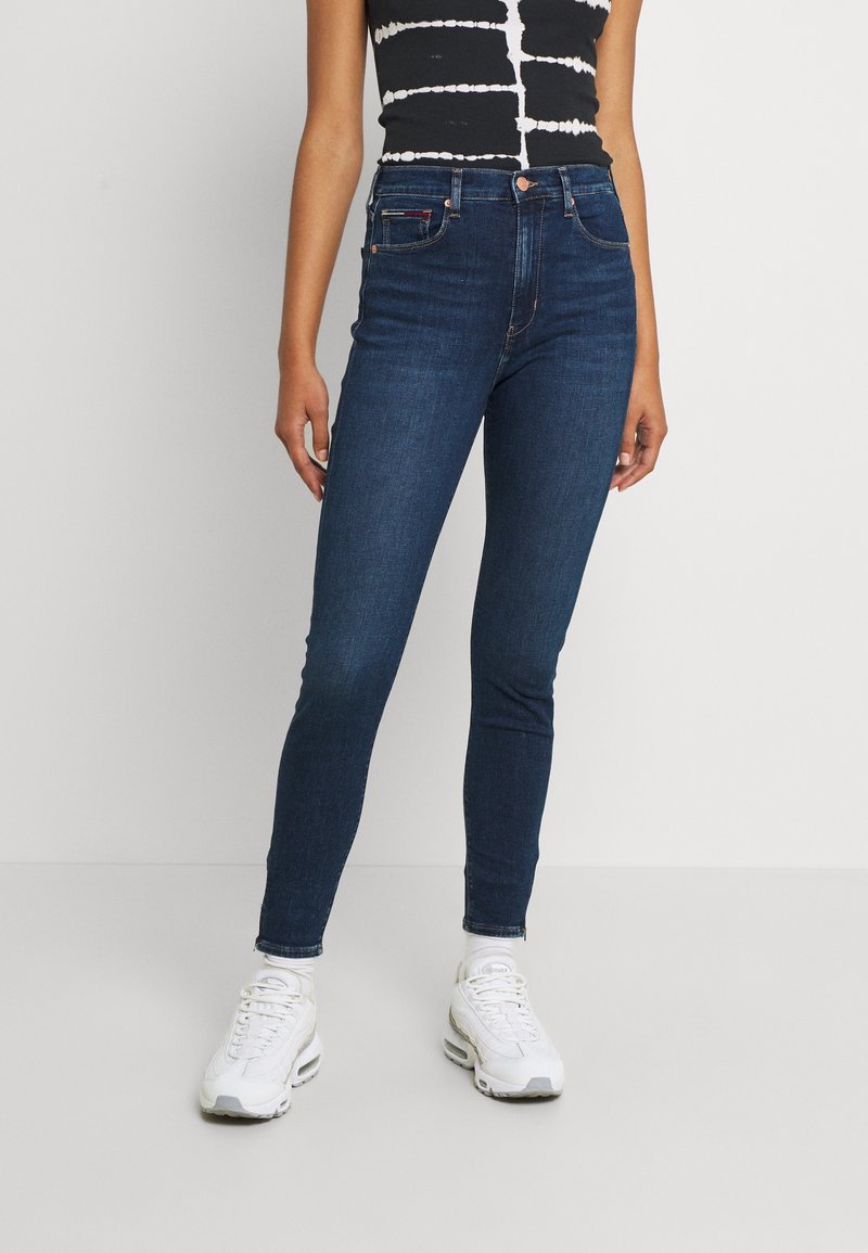 Tmavě modré skinny džíny vyrobené z denimu, s vysokým pasem, pěti kapsami a malou brand tagem vpředu. Nosené s bílými teniskami.