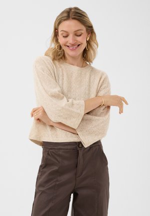 Femme souriante aux cheveux blonds ondulés porte un pull beige texturé, un pantalon marron taille haute et des boucles d’oreilles créoles dorées, bras croisés.