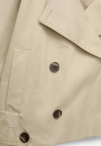 Manteau beige avec des boutons brun foncé, une poche fendue sur le côté gauche et un large col visible en gros plan.