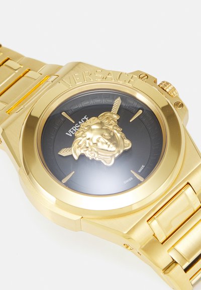 Zelta toņa Versace pulkstenis ar melnu ciparnīcu, izspiestu Medūzas emblēmu un teksturētu siksniņu. Iezīmēts ar iegravētu logo un uzrakstu "Swiss made".