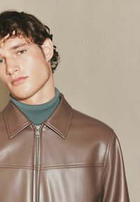 Brun læderjakke med en glat tekstur, der har en spidse krave og frontlynlås. Bæres over en teal turtleneck for kontrast.