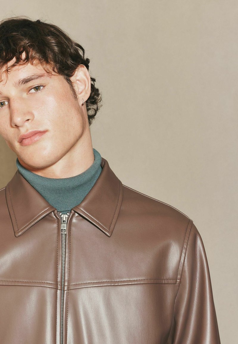 Brun læderjakke med en glat tekstur, der har en spidse krave og frontlynlås. Bæres over en teal turtleneck for kontrast.