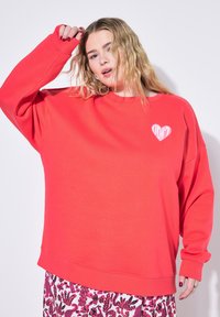 Sweatshirt rouge à col rond avec une coupe ample, arborant un petit graphique en forme de cœur avec le mot "DON'T" en blanc. Tissu doux et poignets côtelés, associé à une jupe à motifs.