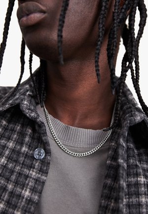 Chemise en plaid gris avec des boutons, superposée sur un pull côtelé gris. Un collier en chaîne argentée repose sur le col, rehaussant le look général.
