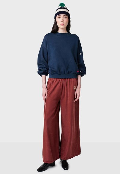 Marinefarbener Oversized-Sweatshirt mit Rundhalsausschnitt, bordeauxfarbene weite Hosen und eine gestreifte Strickmütze mit einem grünen Pompon, kombiniert mit schwarzen Stiefeln.