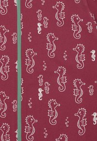 Roter Stoff mit einem wiederholten Muster von weißen Seepferdchen in verschiedenen Größen, umgeben von kleinen Blasen, und einem senkrecht verlaufenden grünen Reißverschluss.