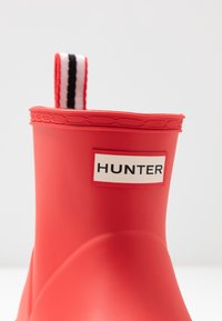 Botte en caoutchouc rouge avec une patte de tirage rayée blanche et noire et une étiquette blanche proéminente avec "HUNTER" en lettres noires.