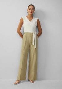 Beige wijde jumpsuit met een aansluitend wit lijfje, V-hals en een tailleband. Model draagt witte sandalen met bandjes en opvallende oorbellen.