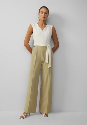 Combinaison ample beige avec un buste ajusté blanc, décolleté en V et ceinture à la taille. Le modèle porte des talons blancs à lanières et des boucles d'oreilles audacieuses.