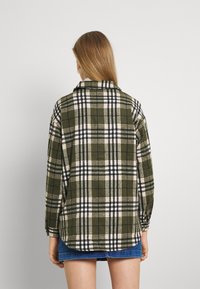 Chemise à carreaux verts avec des carrés noirs, blancs et beiges, dotée d'un col et de manches longues, fabriquée en tissu doux. L'ourlet est légèrement arrondi.