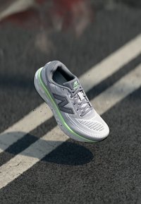 Baskets de course gris clair avec tige en mesh, accents réfléchissants et semelle verte. Comprend un système de laçage et une semelle intermédiaire rembourrée pour le soutien.