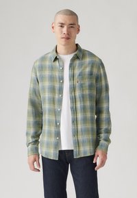 Camisa de franela a cuadros verdes y azules, de mangas largas, con cuello abotonado, un bolsillo en el pecho con logo, usada sobre una camiseta blanca.