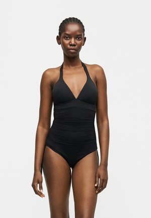 Γυναίκα με κοτσιδωτά μαλλιά που φοράει εφαρμοστό μαύρο ολόσωμο μαγιό με στρας στο λαιμό, στέκεται μπροστά σε απλό ανοιχτό γκρι φόντο.