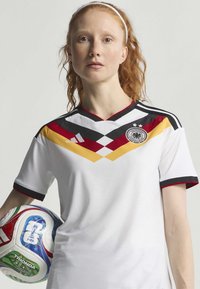 Maillot de football blanc avec des rayures diagonales noires, rouges et jaunes, doté d'un col rond et d'un logo sur la poitrine, tenu avec un ballon de football.