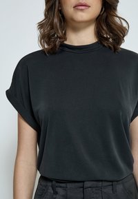 Top corto a maniche corte nero con collo alla coreana, realizzato in tessuto morbido. Presenta maniche arrotolate e una vestibilità rilassata. Abbinato a pantaloni neri.