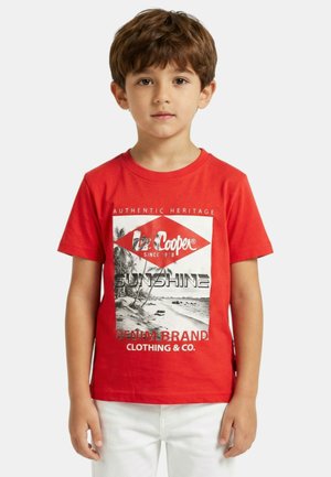 Jeune garçon aux cheveux bruns portant un t-shirt rouge à motif et un pantalon blanc, debout devant un fond clair et uni.