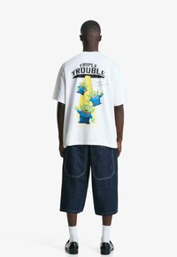 Hvid grafisk t-shirt med tre grønne tegneseriealiens på ryggen, med teksten "TRIPLE TROUBLE" og blå denimshorts.