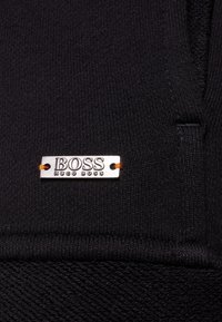 Etichetta metallica con il logo "BOSS HUGO BOSS" cucita su un tessuto di maglia nera con filo arancione.