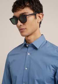 Svijetloplava košulja s dugim rukavima i raširenim ovratnikom, glatke teksture, s bijelim gumbima. Crne sunčane naočale s tamnim staklima upotpunjuju izgled.