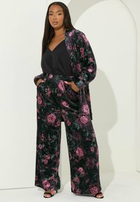Outfit in velluto con una base nera e grandi motivi floreali rosa. La camicia ha maniche lunghe e tasche, abbinata a pantaloni a gamba larga.