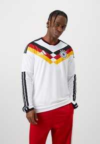 Λευκή μπλούζα με μακρυά μανίκια, με διαγώνιες μαύρες, κόκκινες και κίτρινες λεπτομέρειες, διαθέτει στρογγυλή λαιμόκοψη και μαύρες ρίγες Adidas στα μανίκια.