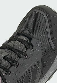 Chaussure de sport noire avec tige en maille et accents texturés, affichant des gouttes d'eau à la surface. Présente un système de laçage et une semelle en caoutchouc.