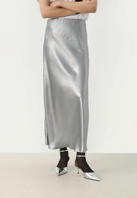 Gonna maxi in satin argento con una texture liscia, dotata di uno spacco laterale e una cintura elastica, indossata con scarpe col tacco argentate a punta.