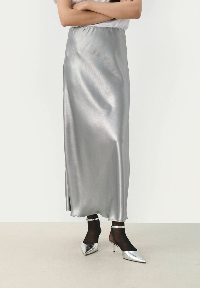 Gonna maxi in satin argento con una texture liscia, dotata di uno spacco laterale e una cintura elastica, indossata con scarpe col tacco argentate a punta.