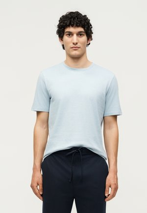 Homme aux cheveux bouclés et foncés portant une chemise bleu clair à manches courtes et un pantalon foncé à cordon, debout devant un fond clair uni.