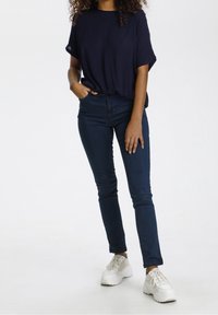 Maglietta corta blu navy, jeans scuri e sneakers bianche. La maglietta ha una vestibilità rilassata; i jeans sono slim con una vita. Le sneakers sono robuste.