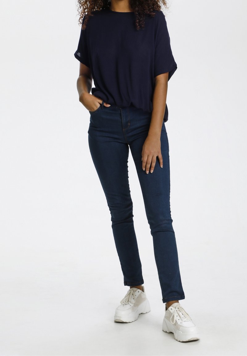Maglietta corta blu navy, jeans scuri e sneakers bianche. La maglietta ha una vestibilità rilassata; i jeans sono slim con una vita. Le sneakers sono robuste.