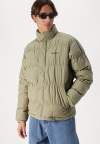 Olivengrønn puffjakke med høy krage, med quiltede seksjoner, brodert logo og frontlommer. Brukt med blå jeans.