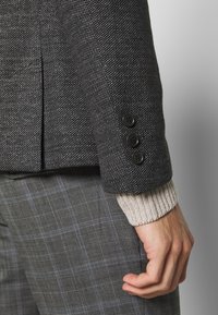 Dunkel strukturierter Blazer mit drei schwarzen Knöpfen am Ärmel, über einem hellen gerippten Pullover und grauen karierten Hosen.