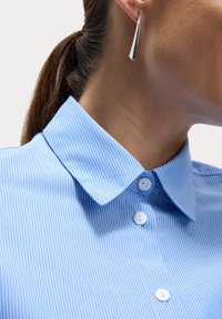 Camicia a righe blu chiaro con colletto, tre bottoni bianchi e una texture in cotone morbido. Orecchino allungato argentato visibile.