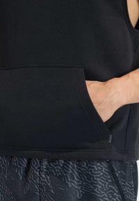 Schwarzer ärmelloser Hoodie mit Fronttasche. Verfügt über eine weiche Textur, ein schlichtes Design und eine lockere Passform. Kombiniert mit gemusterten Shorts.