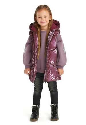 Lila, puffy Weste mit glänzender Oberfläche, getragen über einem helllila Sweatshirt, schwarzer Skinny-Jeans und schwarzen Schnürstiefeln mit Fellfutter.
