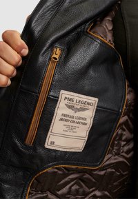 Notranjost črne usnjene jakne z rjavim prešitjem, z zadrgo v temno rjavi barvi ter oznako s podatki o izdelku in blagovno znamko.