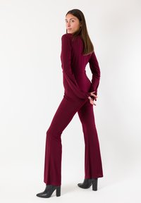 Tuta bordeaux con maniche lunghe e gambe svasate, realizzata in tessuto liscio. Completa l'outfit un paio di stivali neri con tacco alto.