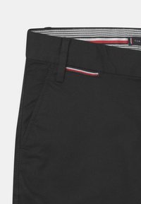 Czarne spodnie z płaskim przodem, z pasem w talii wyściełanym paskowaną podszewką. Zawierają małą kieszonkę z akcentem w czerwono-niebieskiej lamówce.