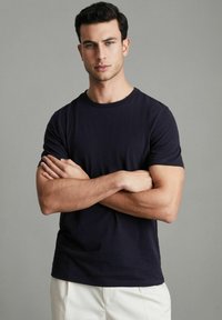 Námornícky modré tričko s okrúhlym výstrihom vyrobené z bavlny. Krátke rukávy, voľný strih a hladká textúra. Model stojí so založenými rukami na šedom pozadí.