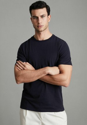 Tumši zili crew neck T-krekli, kas izgatavoti no kokvilnas. Īsas piedurknes, brīva piegriezuma un gluda tekstūra. Modelis stāv ar rokām saliktām krūtīs uz pelēka fona.