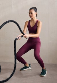 Bordeaux sportski crop top i tajice; crni borbeni užad u rukama; zelene tenisice s bijelim potplatom; glatka betonska pozadina.