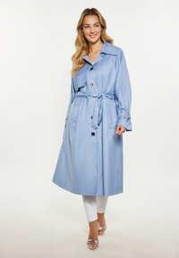 Palton trench de un albastru deschis, realizat dintr-o țesătură netedă, cu un cordon, nasturi mari și un guler clasic, asortat cu pantaloni albi și sandale.
